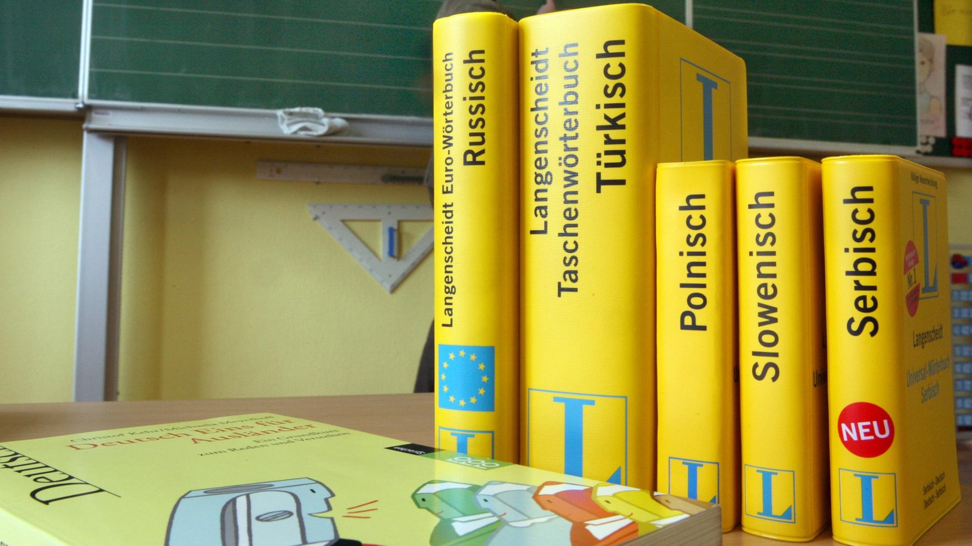 Mehrere Wörterbücher stehen auf einem Tisch in einer Schule. Im Hintergrund ist eine Tafel zu sehen, vor der eine Frau steht. Mehrere Wörterbücher stehen auf einem Tisch in einer Schule. Im Hintergrund ist eine Tafel zu sehen, vor der eine Frau steht.