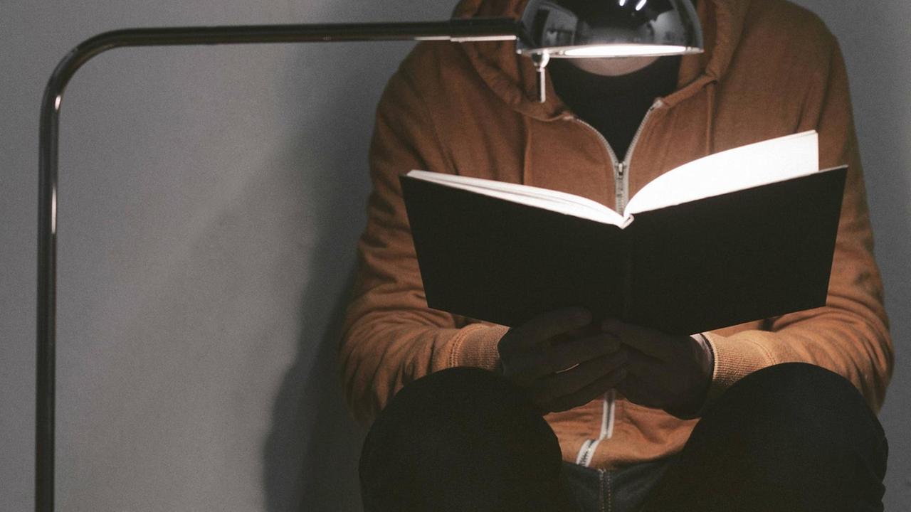 Eine Person hockt unter einer Lampe und liest ein Buch. Eine Person hockt unter einer Lampe und liest ein Buch.