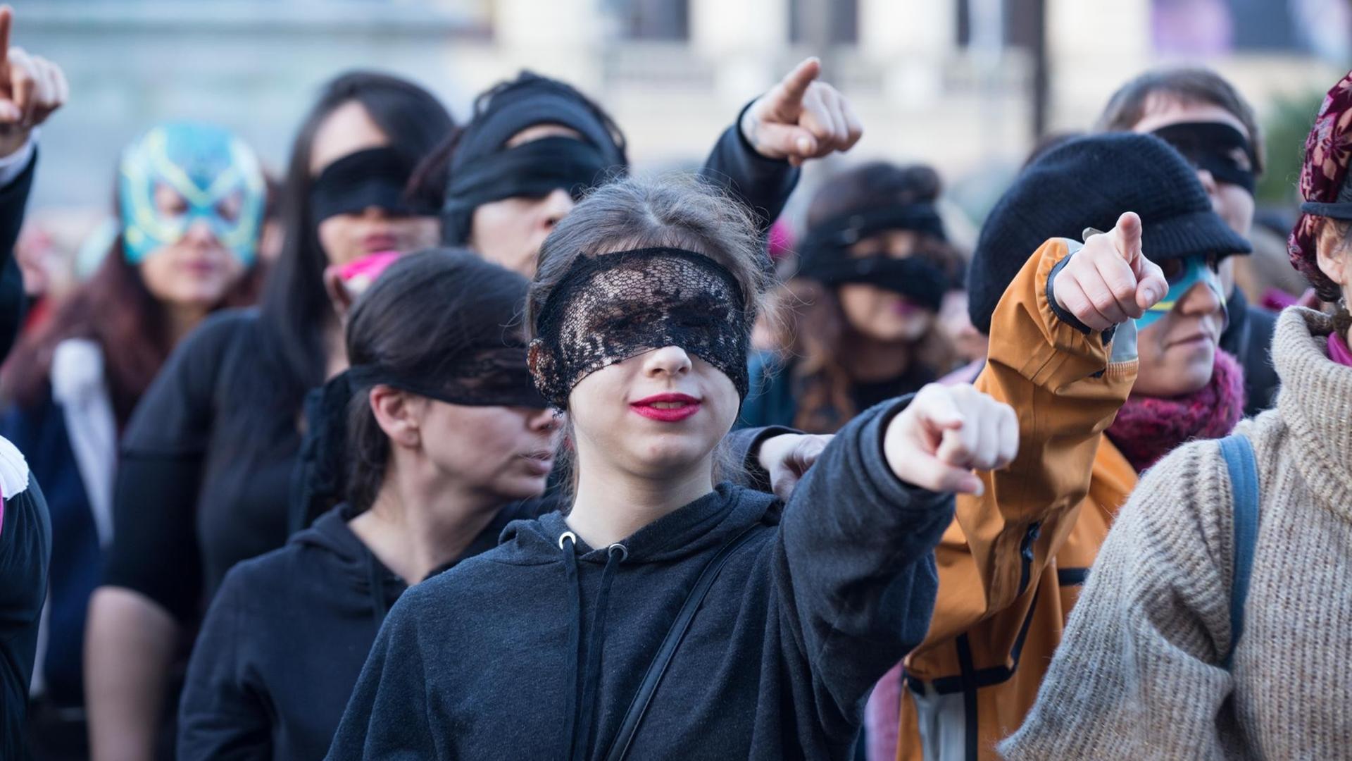 Das Bild zeigt ein Frauenkollektiv, die mit verbundenen Augen, die ausgeschreckten Arme und Zeigefinger auf den Betrachter richten. Das Bild zeigt ein Frauenkollektiv, die mit verbundenen Augen, die ausgeschreckten Arme und Zeigefinger auf den Betrachter richten.