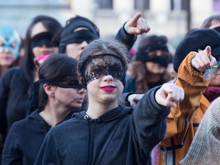 Das Bild zeigt ein Frauenkollektiv, die mit verbundenen Augen, die ausgeschreckten Arme und Zeigefinger auf den Betrachter richten.