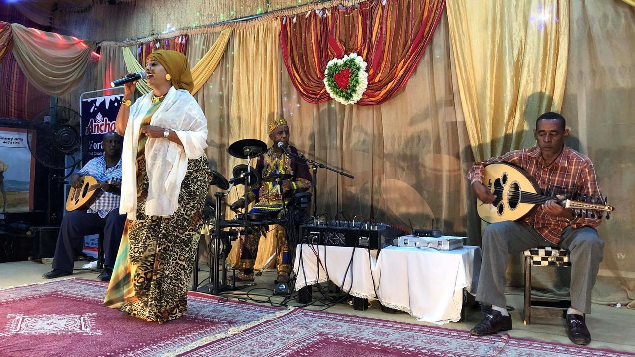 Eine Frau in traditioneller Kleidung steht auf der Bühne, begleitet von 3 Bandmitgliedern: ein Schlagzeuger und zwei Musiker mit Gitarren-ähnlichen Instrumenten. Eine Frau in traditioneller Kleidung steht auf der Bühne, begleitet von 3 Bandmitgliedern: ein Schlagzeuger und zwei Musiker mit Gitarren-ähnlichen Instrumenten.