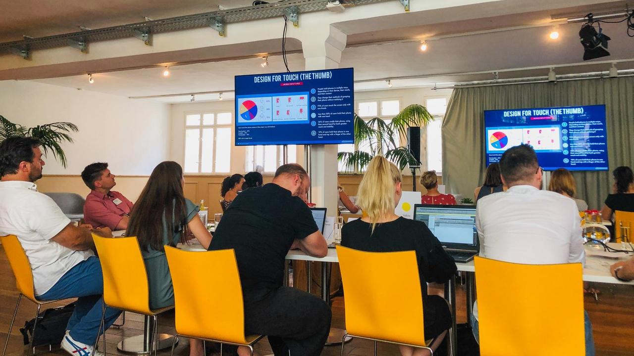 Journalistinnen und sitzen beim "Accelerator-Programm" von Facebook in der Berliner Kalkscheune vor Laptops an Tischen und Blicke auf große Bildschirme, auf der Grafiken gezeigt werden.