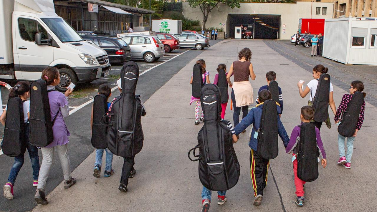 Kleine Kinder, die man von hinten sieht, laufen mit Instrumentenkoffern auf dem Rücken auf das Gebäude des Flughafens Tempelhof zu,