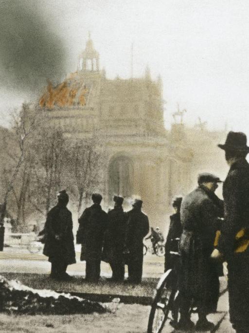 Aufnahme aus dem Februar 1933 im Vordergrund sind Menschen zu sehen, die auf dem im Hintergrund liegenden Reichstag in Berlin blicken, der brennt.