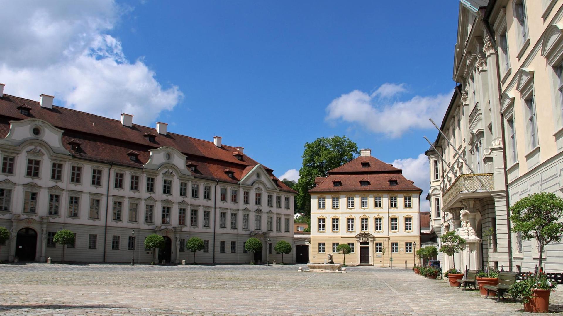 Die Häuser stehen um den gepflasterten rechteckigen Platz mit kleinen Bäumen am Rand. Darüber blauer Himmel mit Wolken. Die Häuser stehen um den gepflasterten rechteckigen Platz mit kleinen Bäumen am Rand. Darüber blauer Himmel mit Wolken.