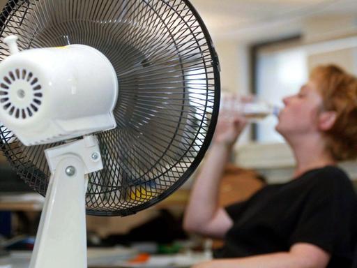 Ein Tisch-Ventilator sorgt in einem Büro in Helsinki für frischen Wind. Im Hintergrund eine Angestellte, die eine tiefen Schluck aus einer Wasserflasche nimmt.