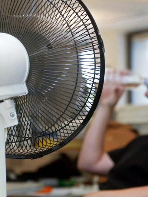 Ein Tisch-Ventilator sorgt in einem Büro in Helsinki für frischen Wind. Im Hintergrund eine Angestellte, die eine tiefen Schluck aus einer Wasserflasche nimmt.
