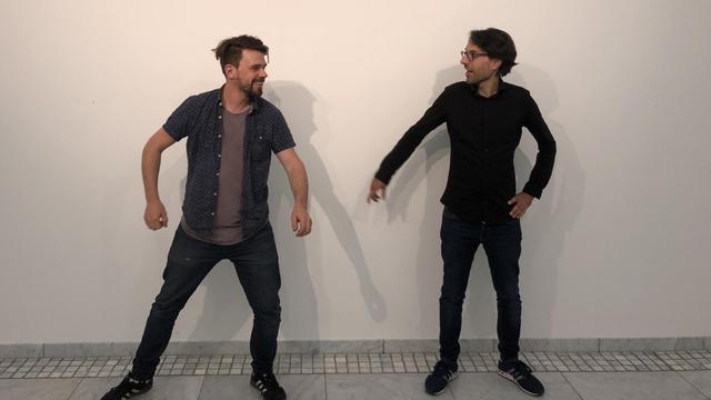 Zwei dunkel gekleidete Männer stehen mit Abstand vor einer weißen Wand, deuten dabei mit ihrer Körperstellung eine sportliche Interaktion an und lachen.
