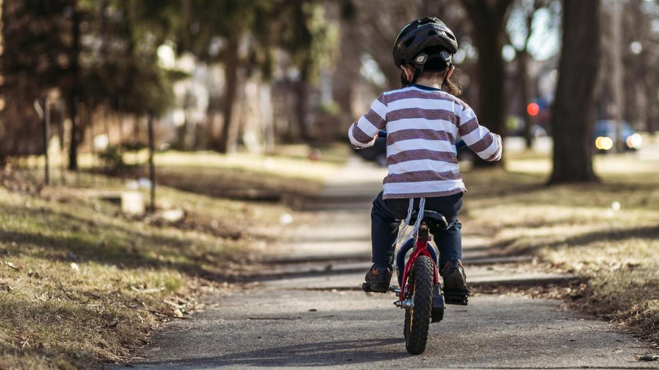 Ein kleines Kind auf einem roten Fahrrad von hinten gesehen. Es fährt einen langen, geraden Fußweg entlang.