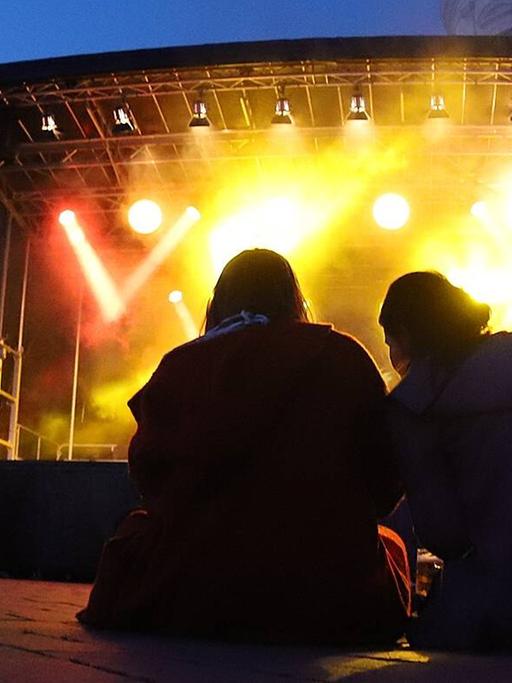 Zwei Menschen sitzen auf dem Boden vor der Open-Air Bühne bei den Musikfestspielen Potsdam Sanssouci. 