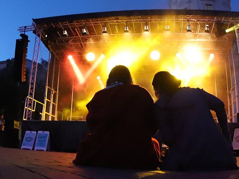 Zwei Menschen sitzen auf dem Boden vor der Open-Air Bühne bei den Musikfestspielen Potsdam Sanssouci. 
