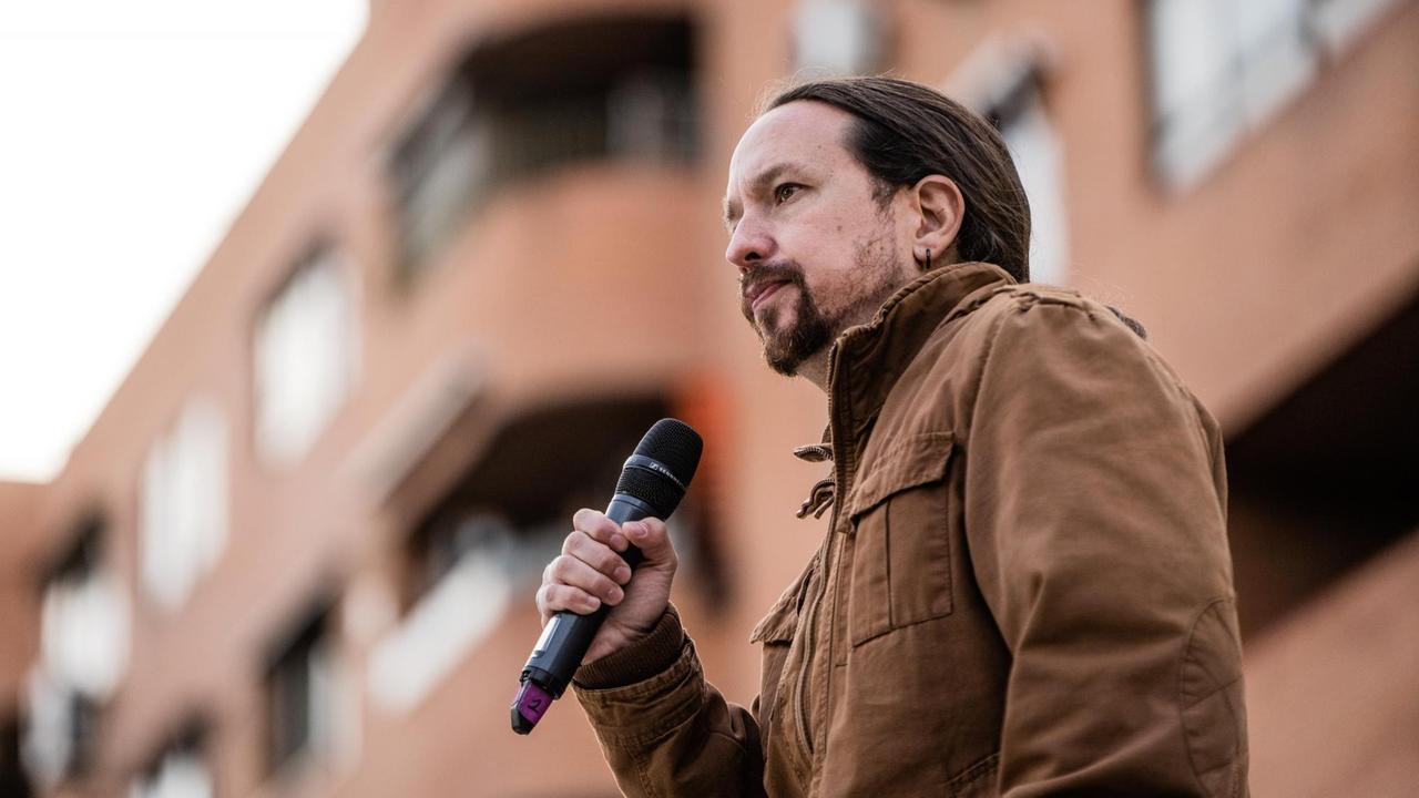 Ein Mann mit Bärtchen und Zopf in einer braunen Jacke steht vor einem Haus mit ernstem Gesichtsausdruck und einem Mikrofon in der Hand.