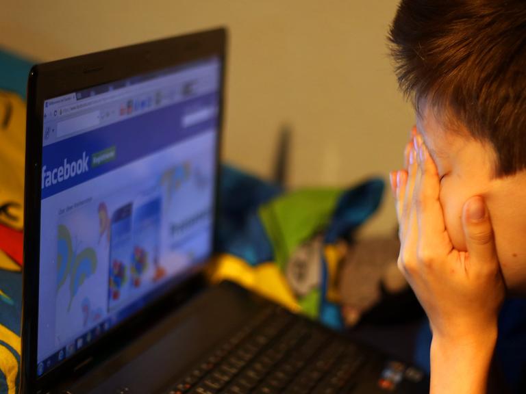 Ein Junge reibt sich vor seinem Laptop beim betrachten der Facebook-Seite die Augen.