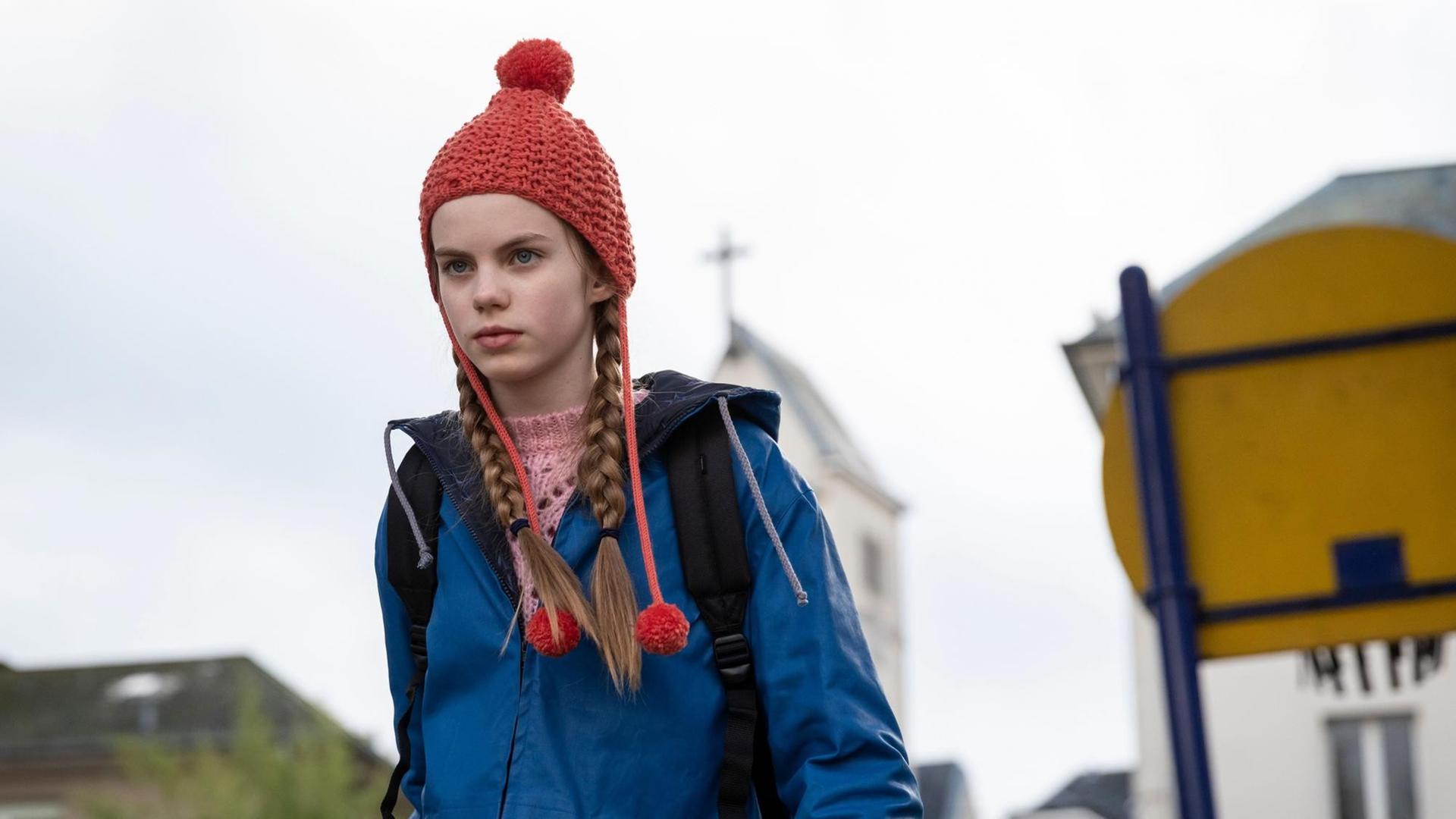 Im Still aus "Mission Ulja Funk" ist Hauptdarstellerin Romy Lou Janinhoff mit einer roten Strickmütze zu sehen, hinter ihr verschwommen die Umrisse einer Kirche. Im Still aus "Mission Ulja Funk" ist Hauptdarstellerin Romy Lou Janinhoff mit einer roten Strickmütze zu sehen, hinter ihr verschwommen die Umrisse einer Kirche.