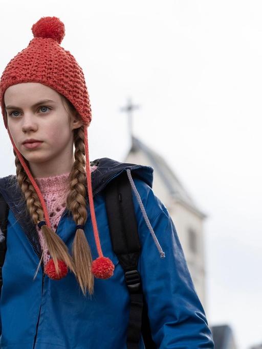 Im Still aus "Mission Ulja Funk" ist Hauptdarstellerin Romy Lou Janinhoff mit einer roten Strickmütze zu sehen, hinter ihr verschwommen die Umrisse einer Kirche.