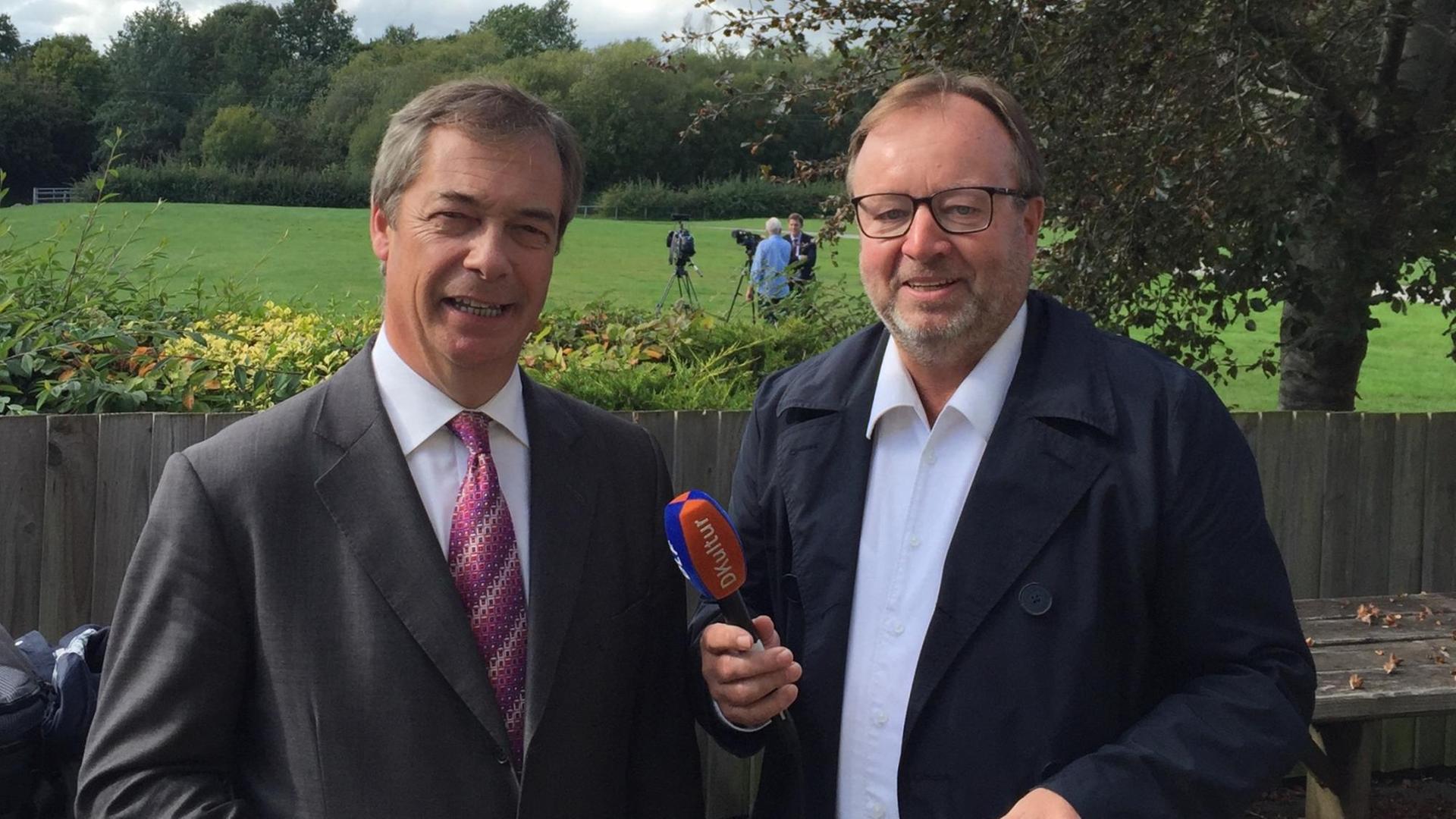 Farage und Meurer stehen lächelnd im Freien nebeneinander und blicken in die Kamera, Meurer hält ein Mikrofon. Hinter ihnen ein Bretterzaun, dahinter eine Wiese, auf der zwei Männer und zwei Kameras auf Stativen stehen. Farage und Meurer stehen lächelnd im Freien nebeneinander und blicken in die Kamera, Meurer hält ein Mikrofon. Hinter ihnen ein Bretterzaun, dahinter eine Wiese, auf der zwei Männer und zwei Kameras auf Stativen stehen.