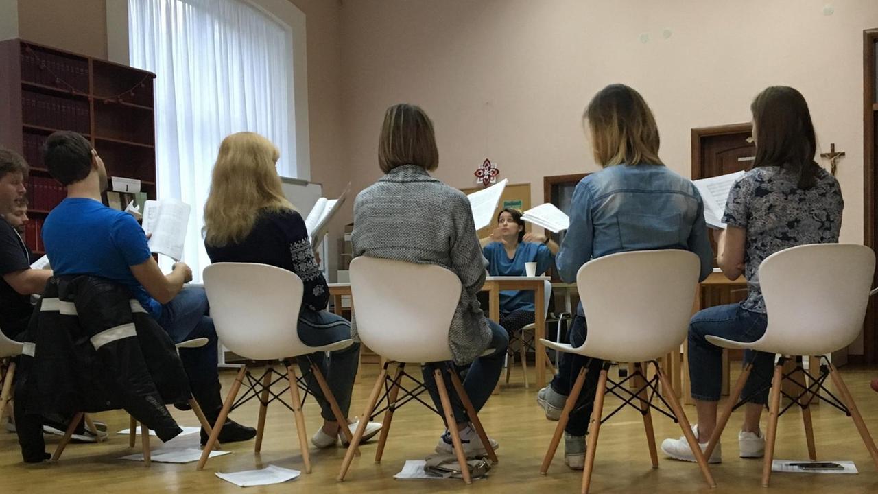 Um eine Frau sitzen mehrere Chormitglieder auf weißen Schalenstühlen und hören ihr zu.