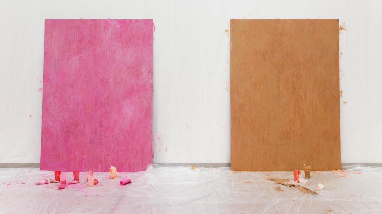 Eine pinke und eine braune Leinwand, gegen eine weiße Wand gelehnt. Davor, auf der Erde, ebenfalls Farbe. Eine pinke und eine braune Leinwand, gegen eine weiße Wand gelehnt. Davor, auf der Erde, ebenfalls Farbe.