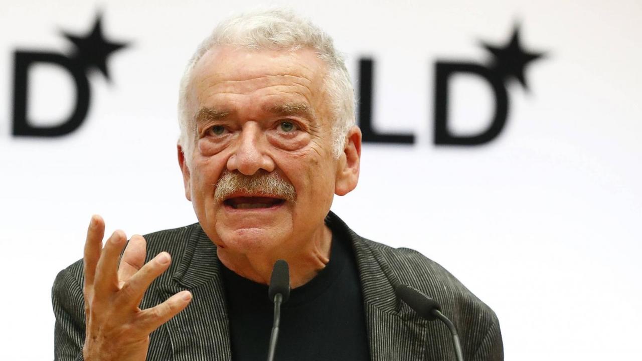 Ein Mann mit kurzem grauen Haar spricht in ein Mikrofon. Dabei gestikuliert er mit seiner rechten Hand. Ein Mann mit kurzem grauen Haar spricht in ein Mikrofon. Dabei gestikuliert er mit seiner rechten Hand.