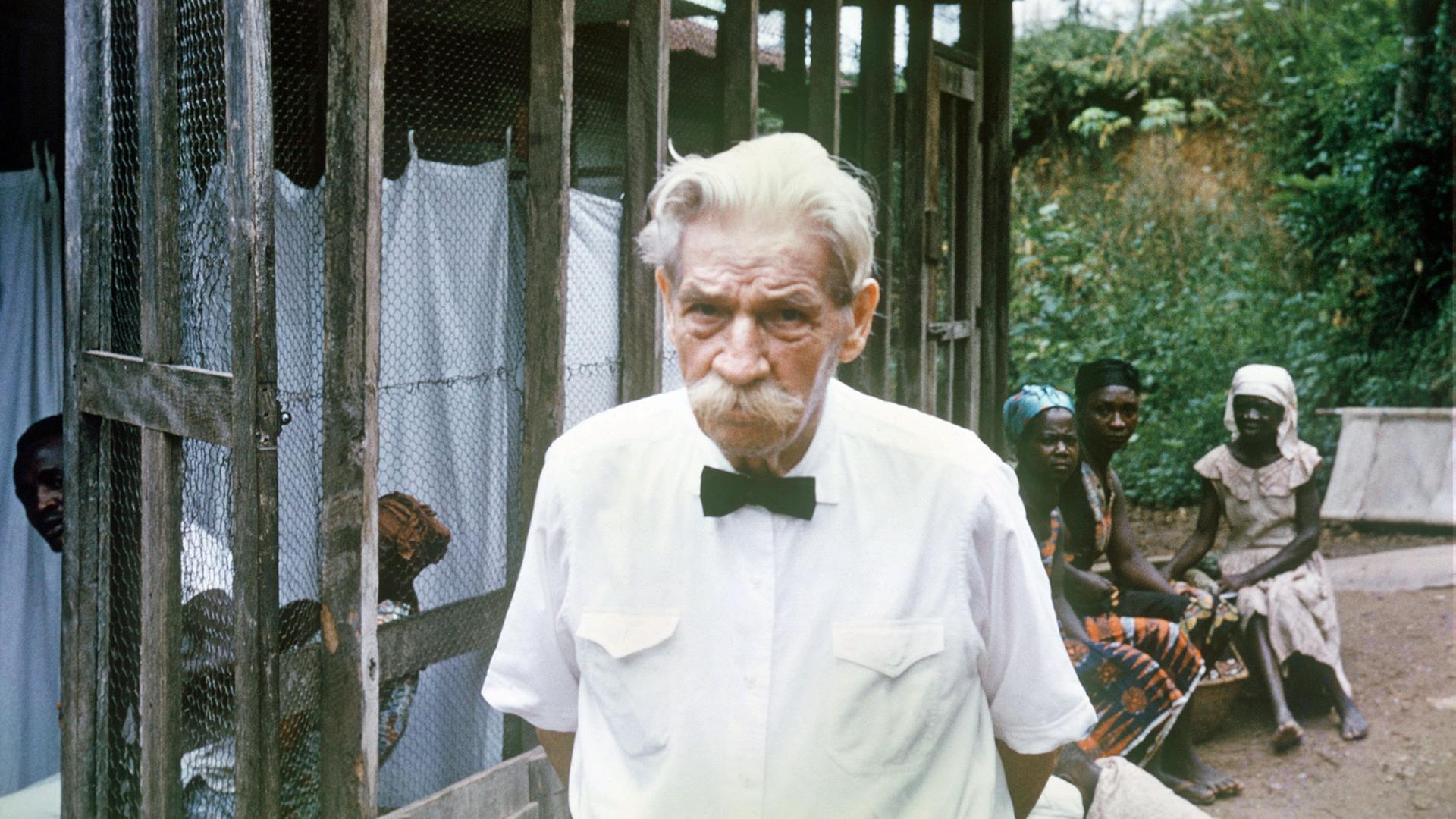 Älterer Mann mit Schnauzbart und einem weißen Hemd in einem afrikanischen Krankenhaus, im Hintergrund sitzen afrikanische Menschen 