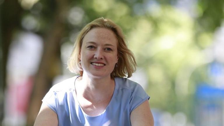 Anika Meenken, Sprecherin des Verkehrsclubs Deutschlands, fährt mit dem Fahrrad durch eine von Bäumen gesäumte Straße und lächelt in die Kamera. Anika Meenken, Sprecherin des Verkehrsclubs Deutschlands, fährt mit dem Fahrrad durch eine von Bäumen gesäumte Straße und lächelt in die Kamera.