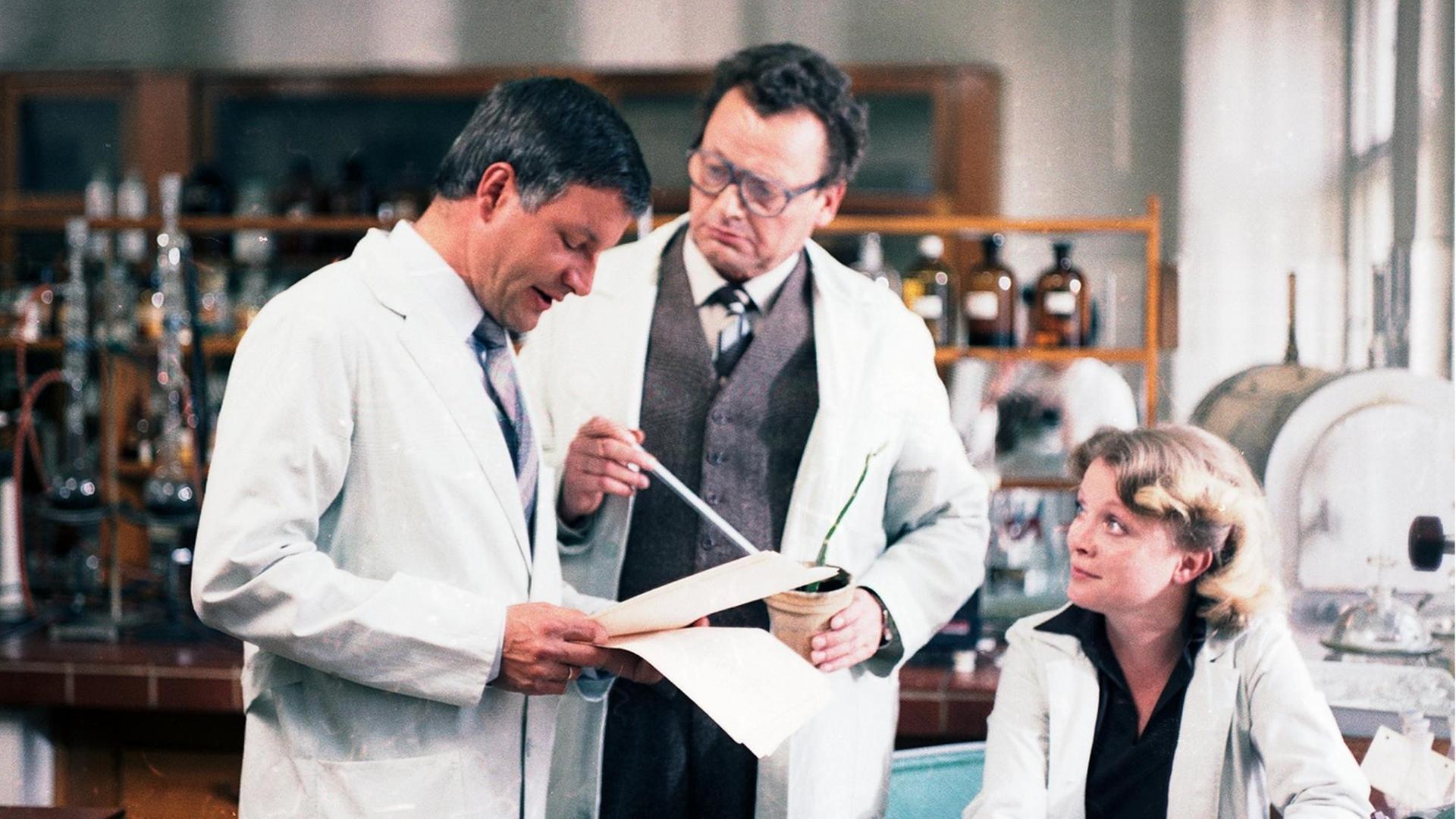 In einem Labor stehen stehen zwei Männer, einer liest in Papieren. Neben ihnen sitzt eine Frau.