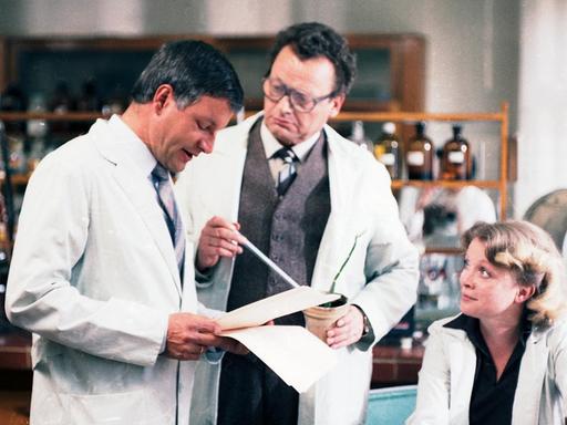 In einem Labor stehen stehen zwei Männer, einer liest in Papieren. Neben ihnen sitzt eine Frau.