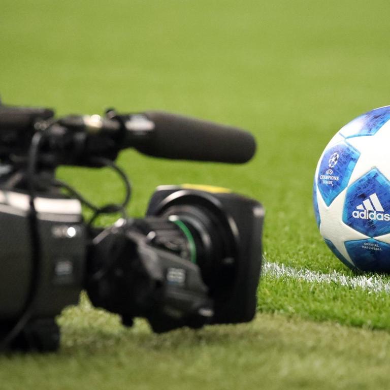 Das Bild zeigt eine Fernsehkamera, die auf dem auf dem Rasen eines Fußballfeldes liegt. Vor der Kamera ist ein Ball zu sehen. 