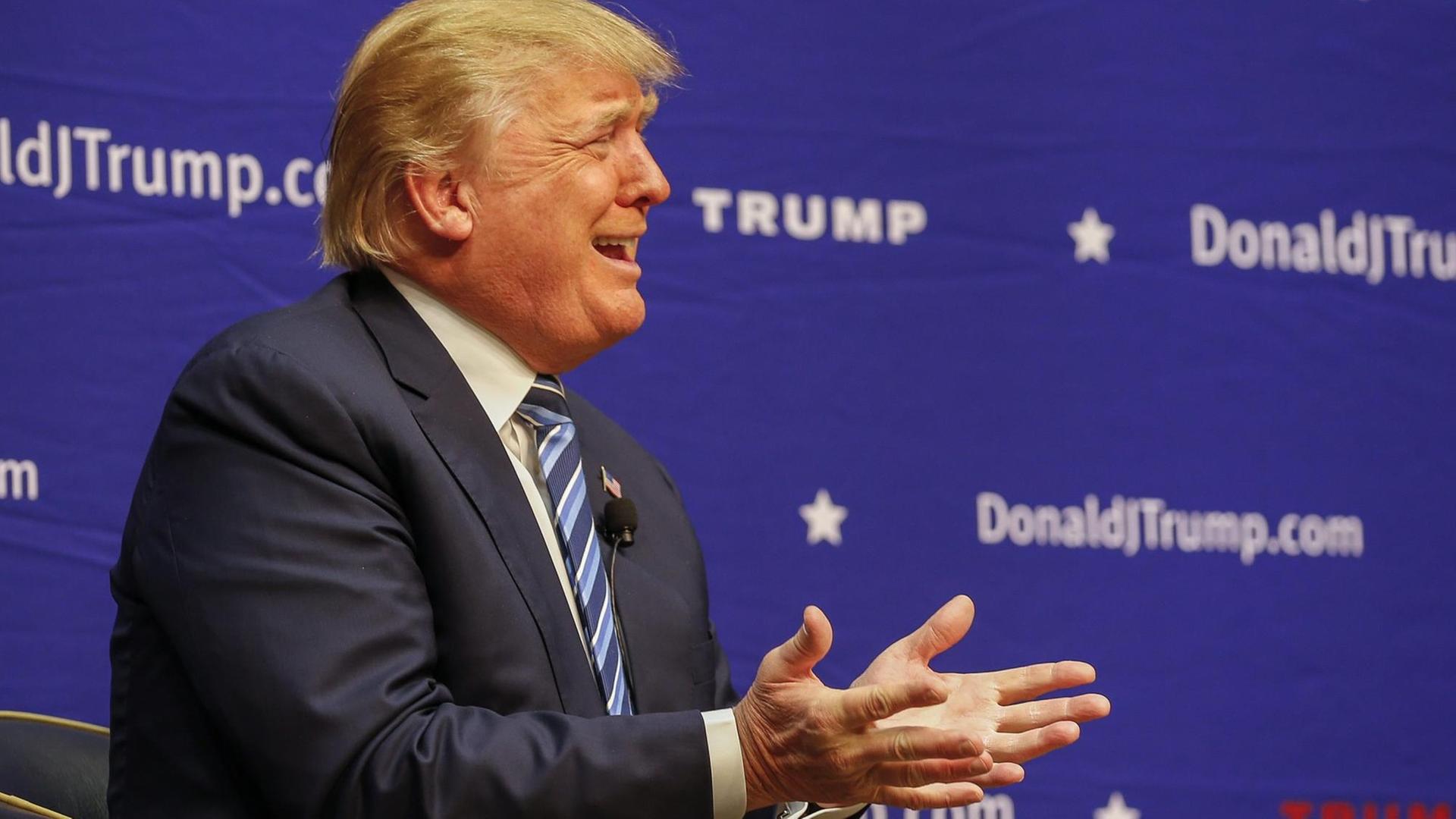 Donald Trump sitzt gestikulierend vor einem blauen Hintergrund mit seinem Namen Donald Trump sitzt gestikulierend vor einem blauen Hintergrund mit seinem Namen