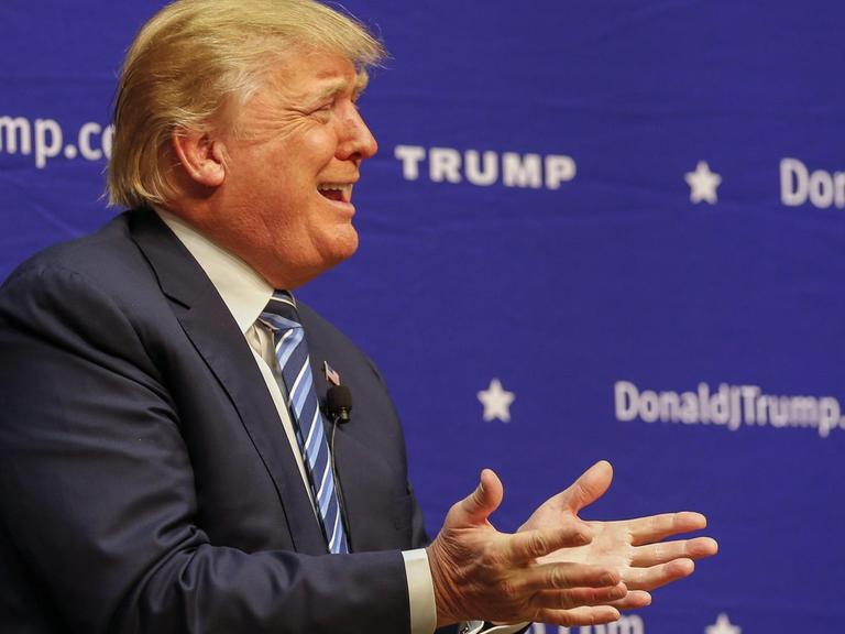 Donald Trump sitzt gestikulierend vor einem blauen Hintergrund mit seinem Namen