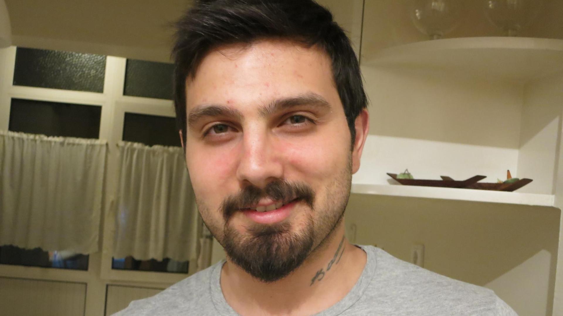 Porträt des türkischen Studenten Melih Bal, schwarze Haare, Drei-Tage-Bart, graues T-Shirt, Tattoo am Hals, lächelnd. Porträt des türkischen Studenten Melih Bal, schwarze Haare, Drei-Tage-Bart, graues T-Shirt, Tattoo am Hals, lächelnd.