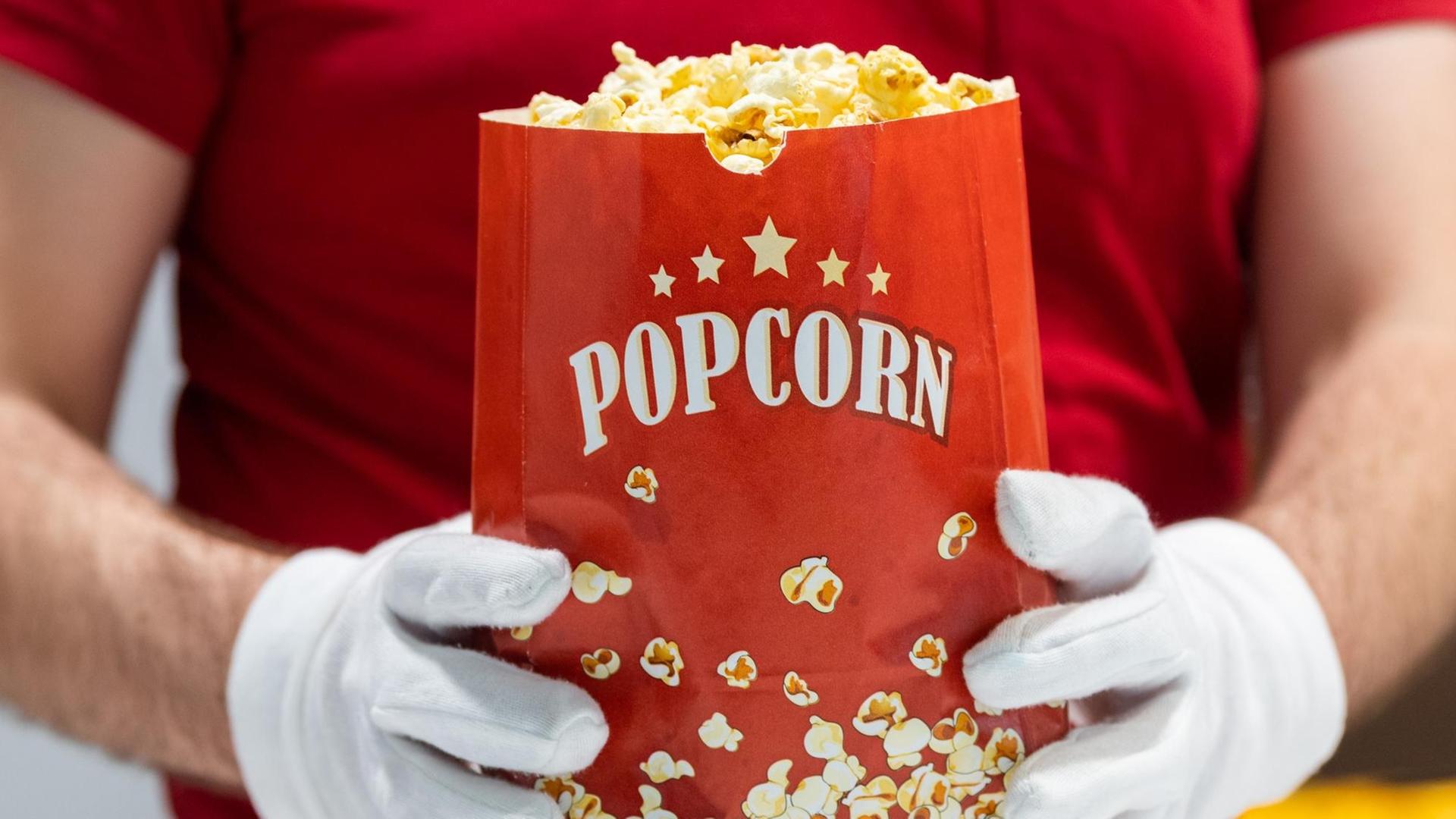 Ein Mitarbeiter des Kinos hält eine Tüte Popcorn in den Händen und trägt dabei Schutzhandschuhe.