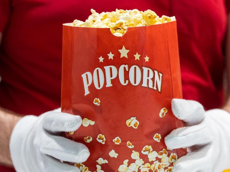 Ein Mitarbeiter des Kinos hält eine Tüte Popcorn in den Händen und trägt dabei Schutzhandschuhe.