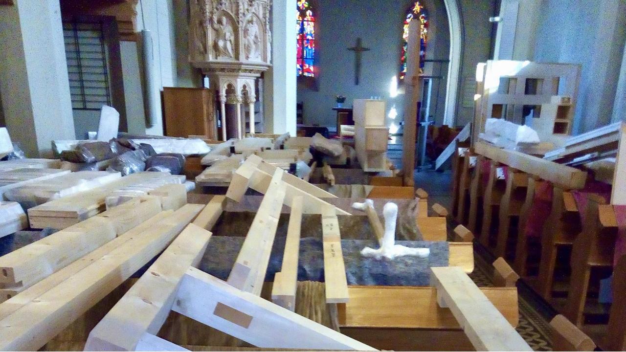 Blick in ein Kirchenschiff, in dem die Sitzbänke als Lagerort für großformatige Holzteile umgenutzt sind. Im Hintergrund sieht man ein Kreuz, die Kanzel und Kirchenfenster.