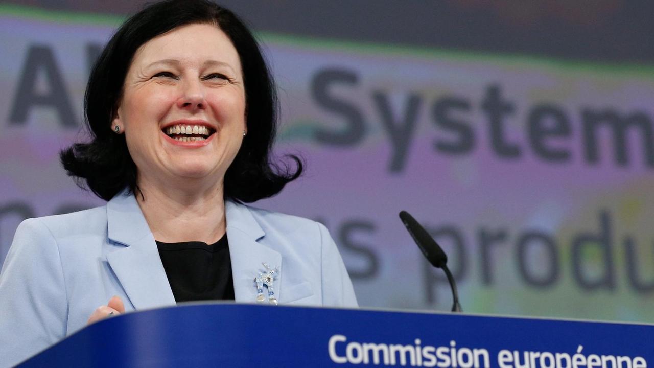 Jourova steht lachend an einem Rednerpult mit Mikrofon und der Aufschrift "Commission européennne - European Commission".