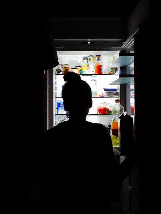 Silhouette einer Frau nachts vor dem offenen Kühlschrank.
