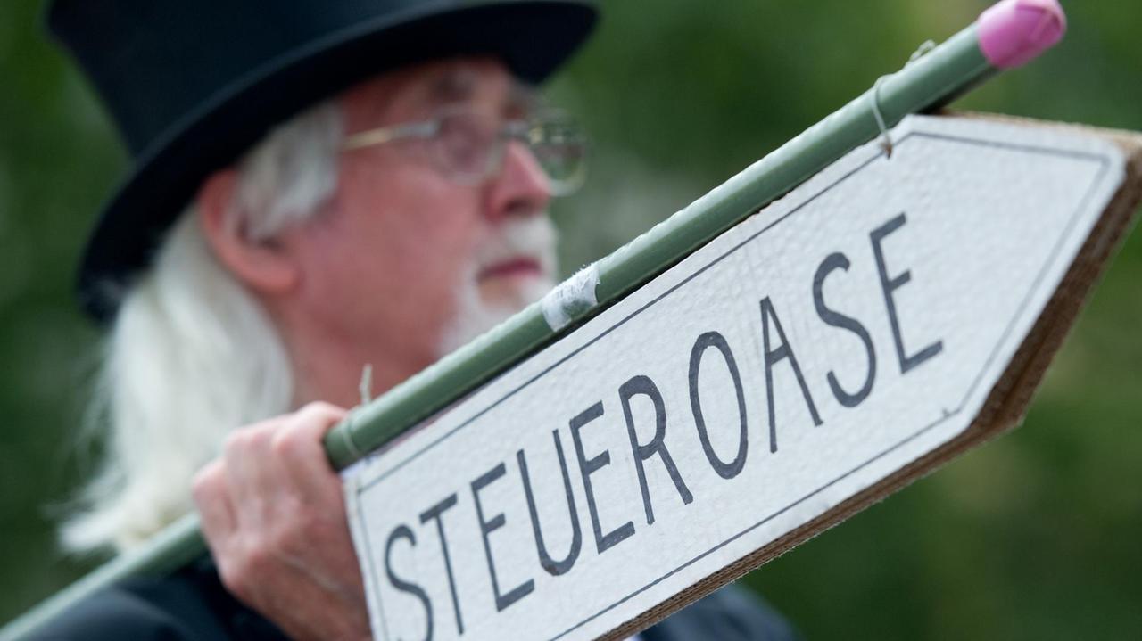 Der ältere weißhaarige Demonstrant mit Bart trägt einen Zylinder und hält ein Richtungs-Schild mit der Aufschrift "Steueroase" in der Hand. Das Schild ist scharf im Vordergrund, der Kopf des Mannes unscharf. Der ältere weißhaarige Demonstrant mit Bart trägt einen Zylinder und hält ein Richtungs-Schild mit der Aufschrift "Steueroase" in der Hand. Das Schild ist scharf im Vordergrund, der Kopf des Mannes unscharf.