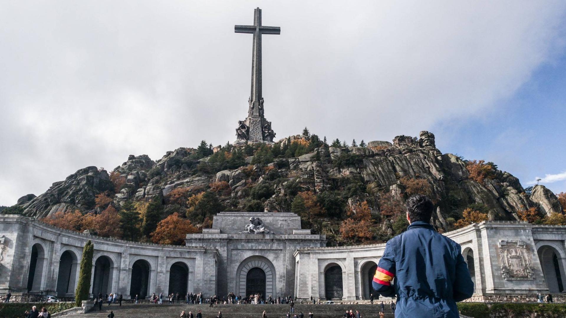 Ein Mann mit einer spanischen Armbinde und einer blauen Jacke steht vor dem "Tal der Gefallenen" und sieht zum riesigen Kreuz in der Mitte des Berges. Ein Mann mit einer spanischen Armbinde und einer blauen Jacke steht vor dem "Tal der Gefallenen" und sieht zum riesigen Kreuz in der Mitte des Berges.