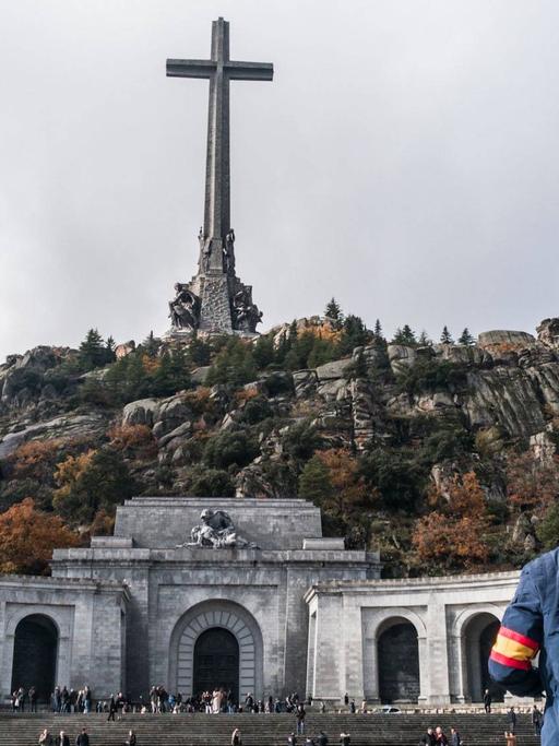 Ein Mann mit einer spanischen Armbinde und einer blauen Jacke steht vor dem "Tal der Gefallenen" und sieht zum riesigen Kreuz in der Mitte des Berges.