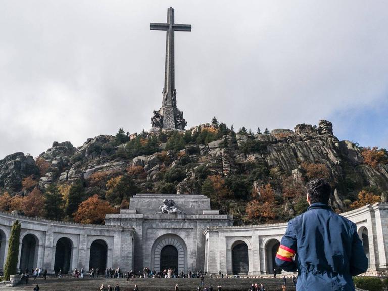 Ein Mann mit einer spanischen Armbinde und einer blauen Jacke steht vor dem "Tal der Gefallenen" und sieht zum riesigen Kreuz in der Mitte des Berges.