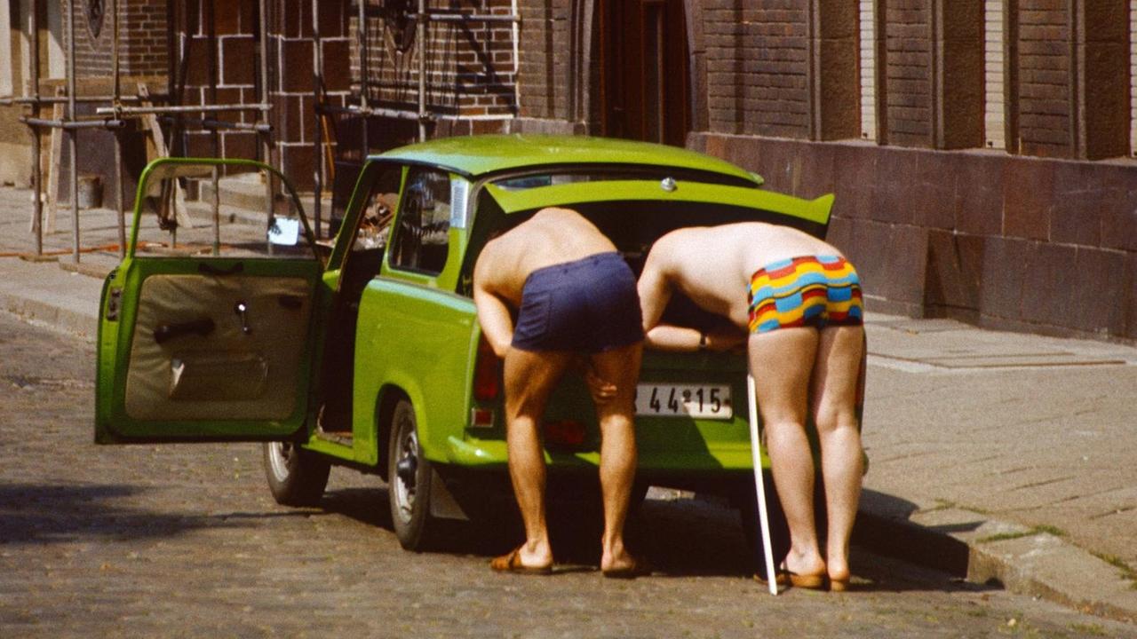 Zwei ältere Herren in farbenfrohen Badehosen beugen sich in den Kofferraum eines Trabants. Aufgenommen 1985 in Berlin.