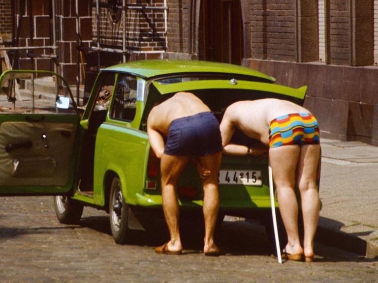 Zwei ältere Herren in farbenfrohen Badehosen beugen sich in den Kofferraum eines Trabants. Aufgenommen 1985 in Berlin.