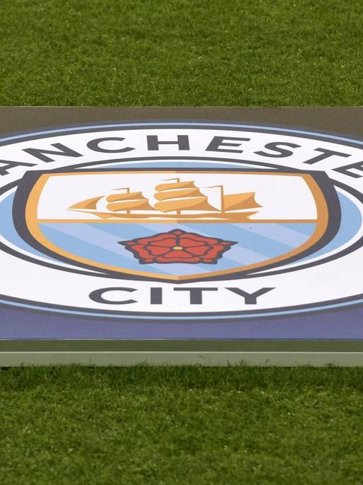 Das Bild zeigt das Logo des englischen Fußballvereins Manchester City. Es liegt im Stadion ausgebreitet auf dem Rasen.