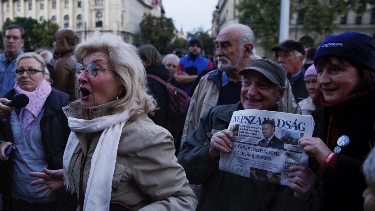 Mehrere Menschen vor dem Parlamentsgebäude in Ungarn, eine Frau ruft laut etwas, zwei halten eine Ausgabe der Zeitung Nepszabadsag. Mehrere Menschen vor dem Parlamentsgebäude in Ungarn, eine Frau ruft laut etwas, zwei halten eine Ausgabe der Zeitung Nepszabadsag.