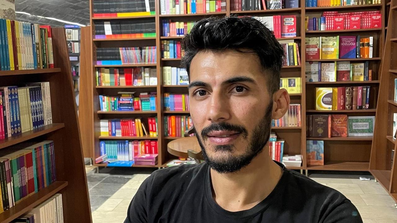 Ein junger Mann mit schwarzem T-Shirt sitzt vor vollen Bücherregalen. Ein junger Mann mit schwarzem T-Shirt sitzt vor vollen Bücherregalen.