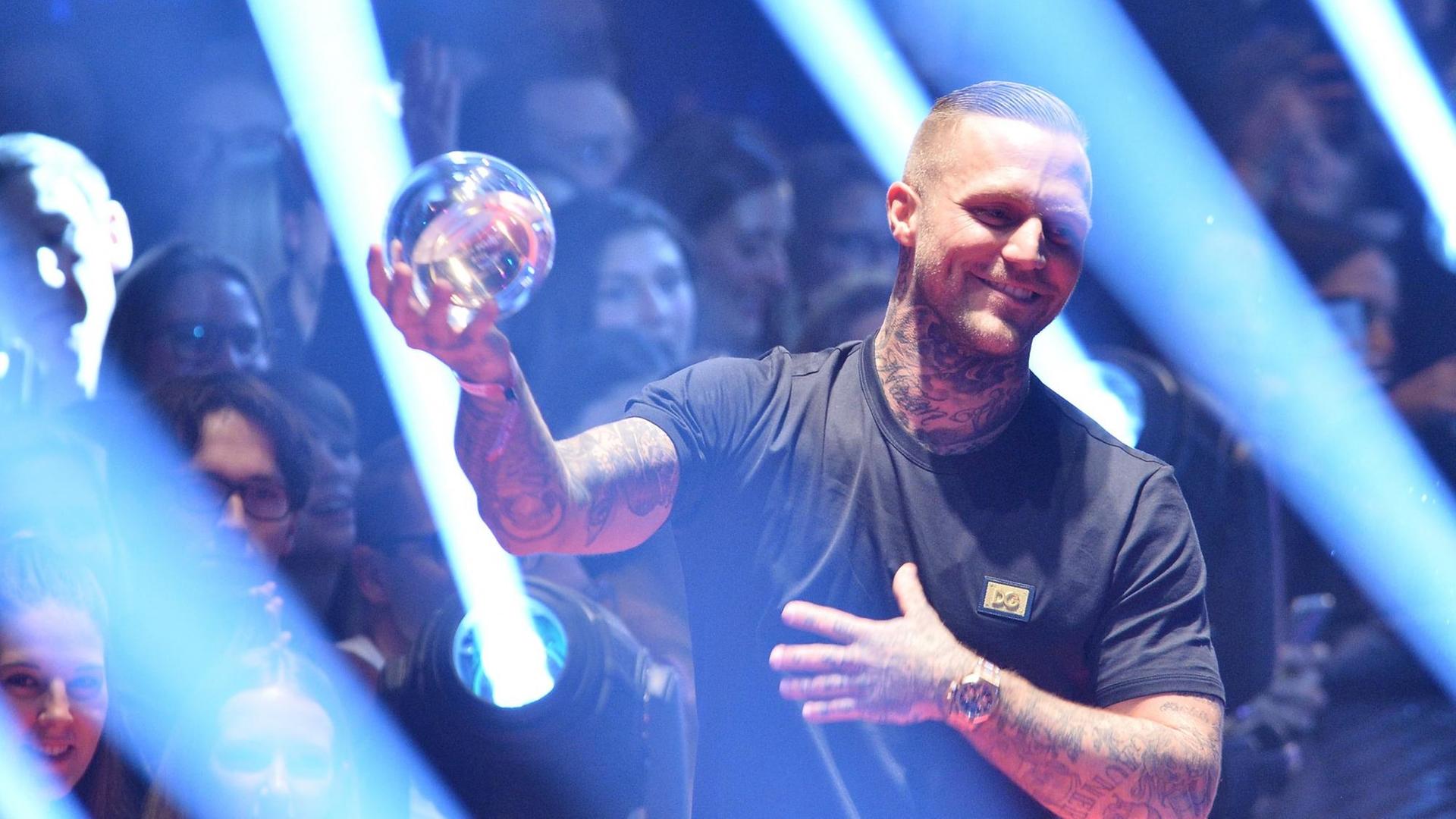 Kontra K steht im Lichtspiel auf der Bühne und zeigt den Pokal, eine Glaskugel. Kontra K steht im Lichtspiel auf der Bühne und zeigt den Pokal, eine Glaskugel.