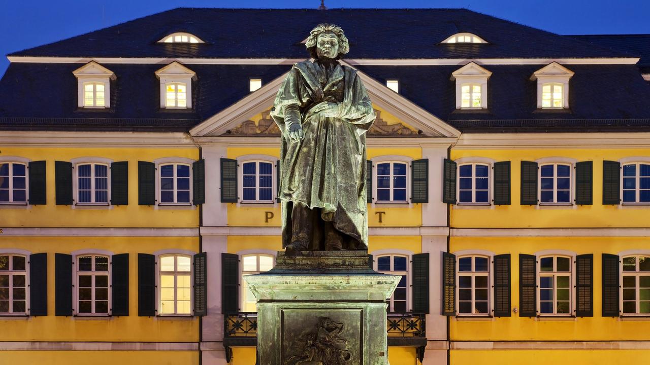 Im Vordergrund das Beethoven-Denkmal, im Hintergrund das Hauptpostamt Bonns. Das Denkmal besteht aus einer überlebensgroßen Darstellung Beethovens auf einem Sockel, das hier am Abend angestrahlt wird und sich daher deutlich von dem gelb gestrichenen mehrstöckigen Gebäude im Hintergrund abhebt.