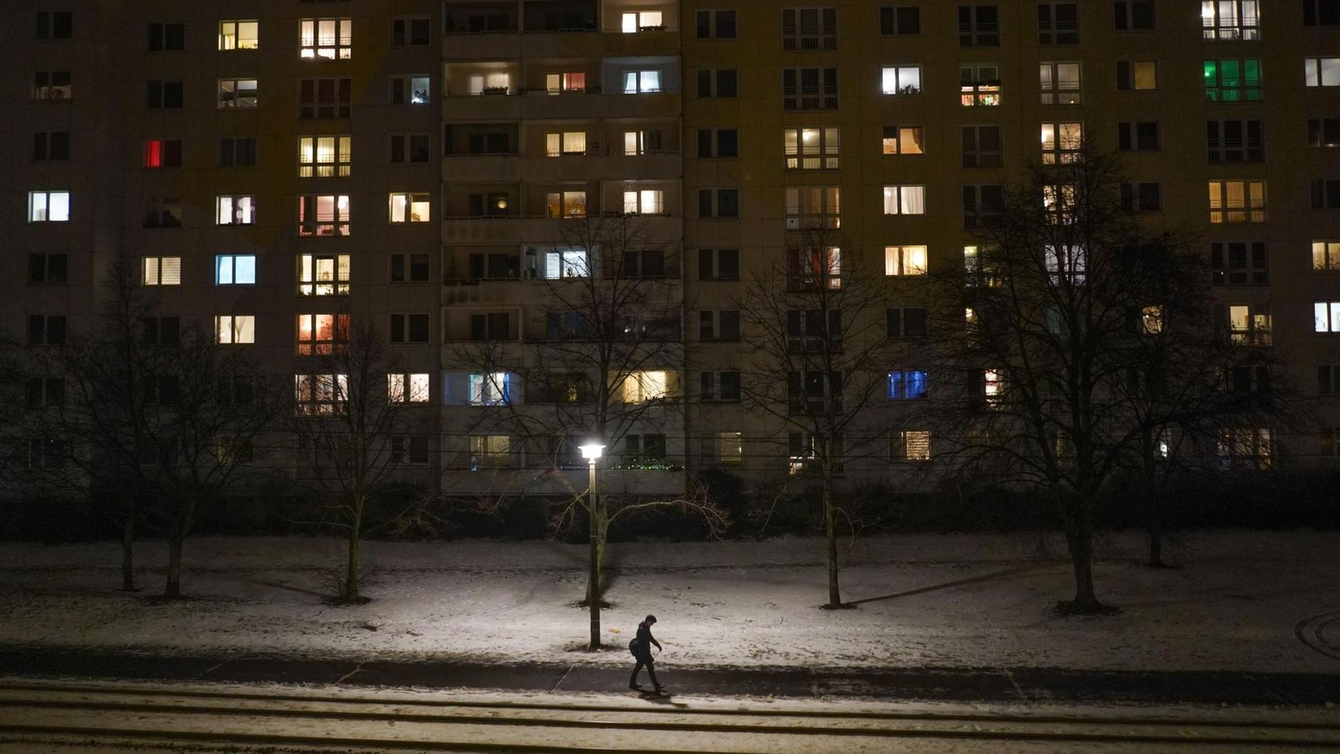 Teilweise hell erleuchtet sind einige Zimmer eines Mehrfamilienhauses am Abend – ein Mann geht allein die Straße entlang. Teilweise hell erleuchtet sind einige Zimmer eines Mehrfamilienhauses am Abend – ein Mann geht allein die Straße entlang.