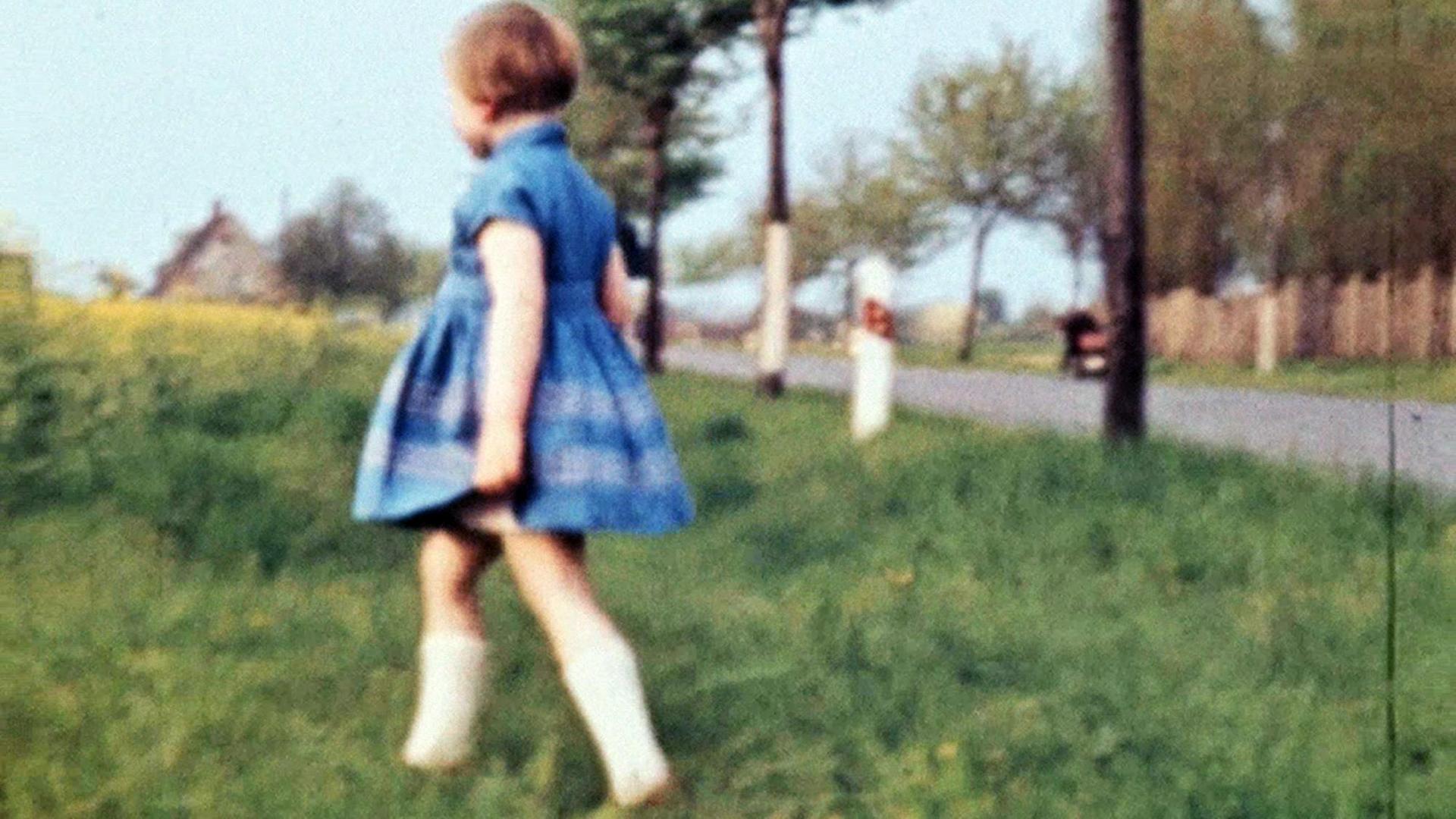 Ein Mädchen in einem Kleid läuft über eine Wiese, im Hintergrund ist eine Allee zu sehen, typisch körnige DDR-Film_Aufnahme Ein Mädchen in einem Kleid läuft über eine Wiese, im Hintergrund ist eine Allee zu sehen, typisch körnige DDR-Film_Aufnahme