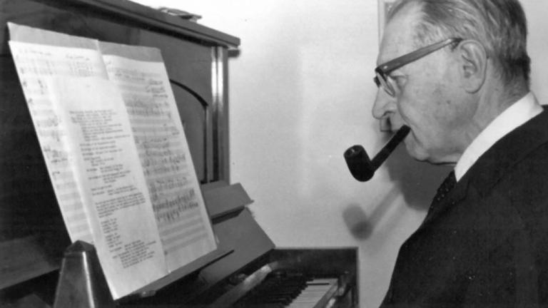 Ein älterer Mann mit Pfeife sitzt an einem Klavier und spielt aus großformatigen Notenblättern.